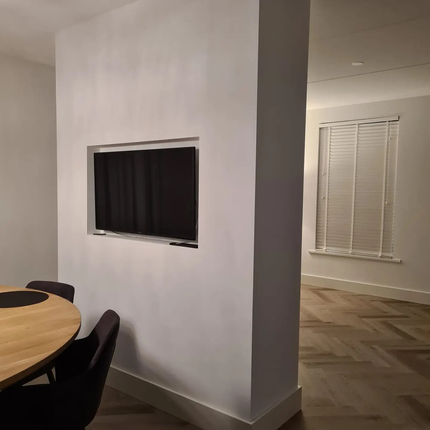 Glad gestuct tussenmuur met inbouwnis voor de tv, naast een eikenhouten tafel en een visgraatvloer